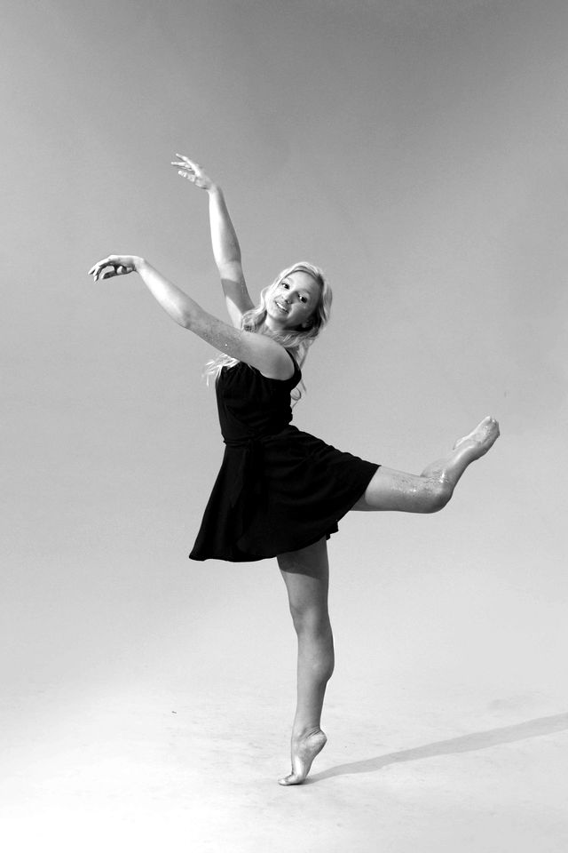 A dancer in a black dress balancing on one leg against a tan backdrop