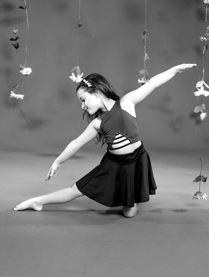 A dancer in a green top and brown skirt in a long kneeling lunge, one arm raised, against a rust backdrop hung with flowers