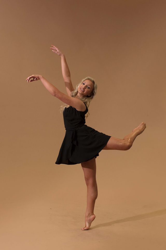 A dancer in a black dress balancing on one leg against a tan backdrop