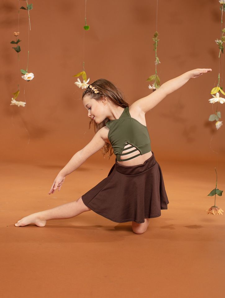 A dancer in a green top and brown skirt in a long kneeling lunge, one arm raised, against a rust backdrop hung with flowers